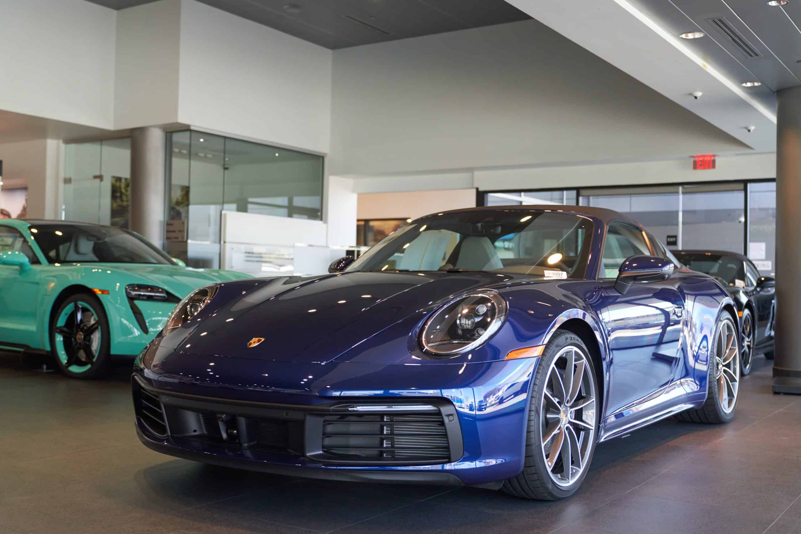 A dark blue Porsche 911 sports car is prominently featured in the foreground of a brightly lit dealership showroom. Behind it, a light teal Porsche Taycan electric sedan is visible to the left, and another dark car is partially seen further back on the right. The showroom has reflective floors and glass walls, creating a sleek, modern environment.