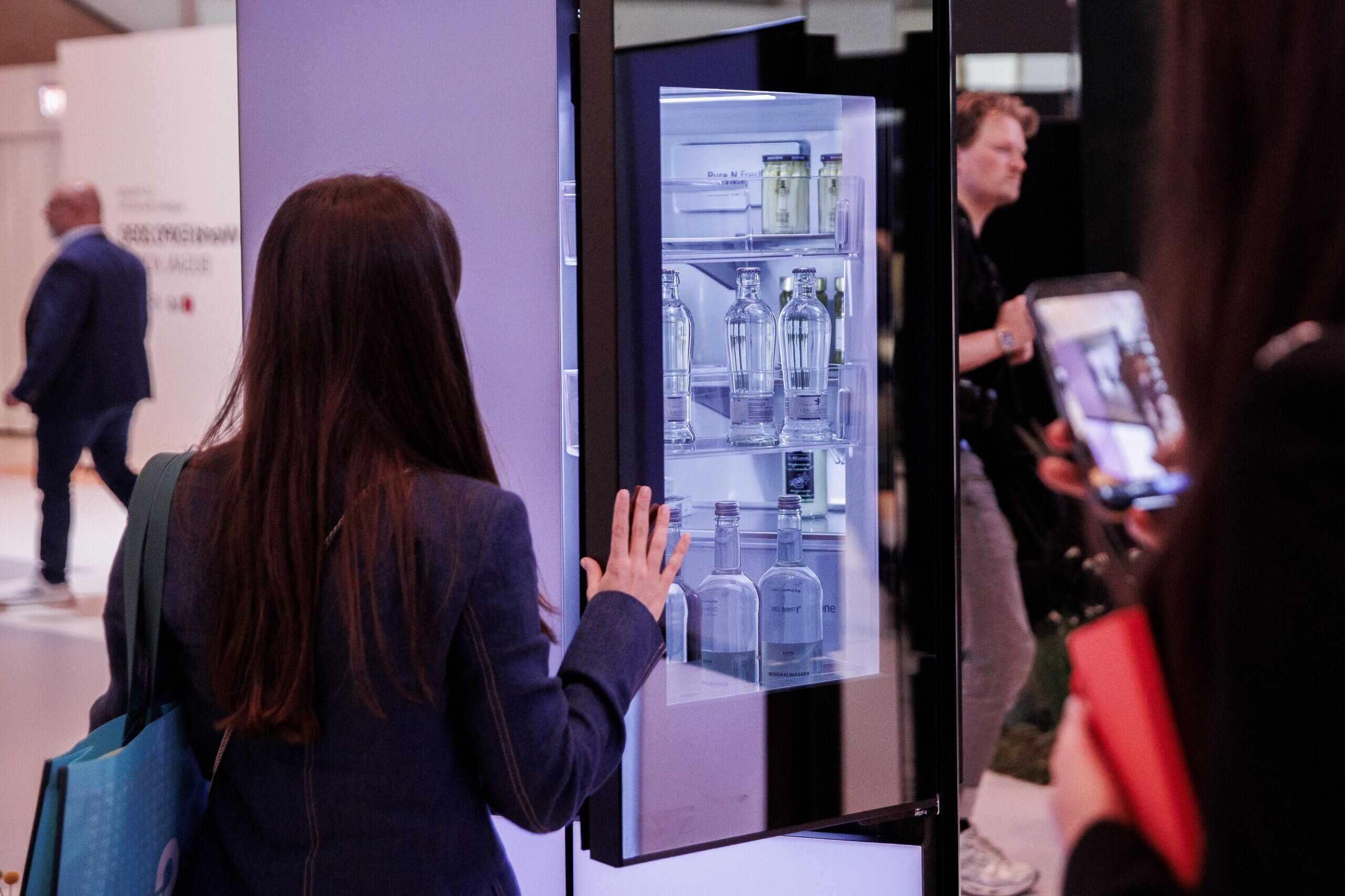 A person with long brown hair and a dark blazer is seen from behind, reaching with their right hand towards an open, brightly lit refrigerator or display cabinet. The cabinet features clear glass shelves holding rows of clear glass bottles, some with white labels. In the blurred background, other people are visible, one holding a smartphone to take a picture, and another man in a suit walking away.
