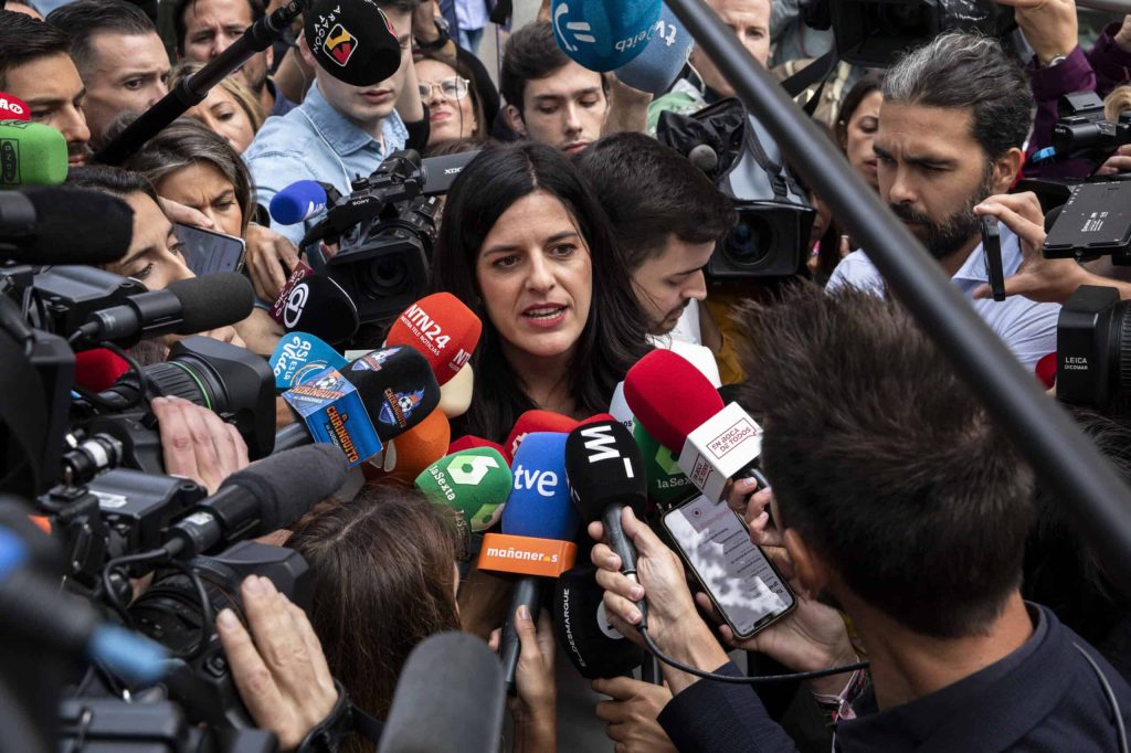 A woman with dark hair and a serious expression is surrounded by dozens of colorful microphones from various news organizations and several camera lenses, all pointed at her. She is speaking, her mouth slightly open, amidst a throng of reporters and camera operators in a crowded, high-pressure media scrum.