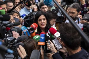 A woman with dark hair and a serious expression is surrounded by dozens of colorful microphones from various news organizations and several camera lenses, all pointed at her. She is speaking, her mouth slightly open, amidst a throng of reporters and camera operators in a crowded, high-pressure media scrum.