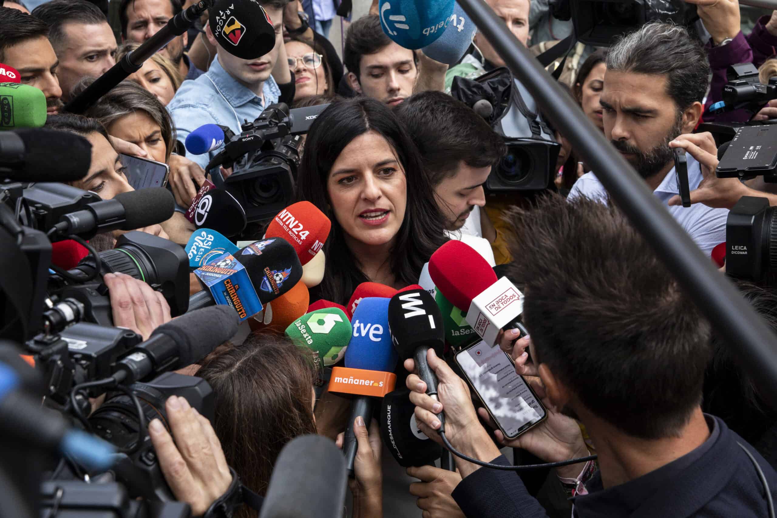 A woman with dark hair and a serious expression is surrounded by dozens of colorful microphones from various news organizations and several camera lenses, all pointed at her. She is speaking, her mouth slightly open, amidst a throng of reporters and camera operators in a crowded, high-pressure media scrum.