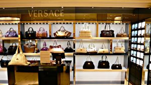 A wide shot of a luxury handbag display inside a high-end store. Numerous designer handbags in various colors like purple, black, brown, cream, and metallic gold are arranged on illuminated shelves. The brand name 'VERSACE' is visible in gold letters above the display. The lighting is warm, highlighting the polished surfaces of the bags and the store's opulent ambiance.