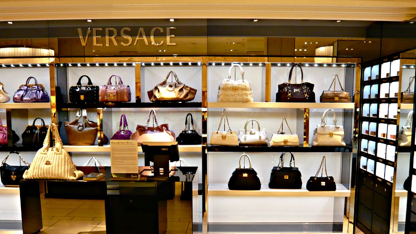 A wide shot of a luxury handbag display inside a high-end store. Numerous designer handbags in various colors like purple, black, brown, cream, and metallic gold are arranged on illuminated shelves. The brand name 'VERSACE' is visible in gold letters above the display. The lighting is warm, highlighting the polished surfaces of the bags and the store's opulent ambiance.