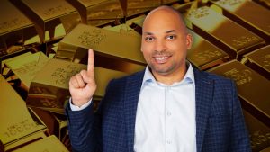 A smiling bald man in a blue plaid suit jacket and light blue collared shirt points his right index finger upwards. He is positioned in the foreground against a blurred background of numerous stacked, shiny gold bars.