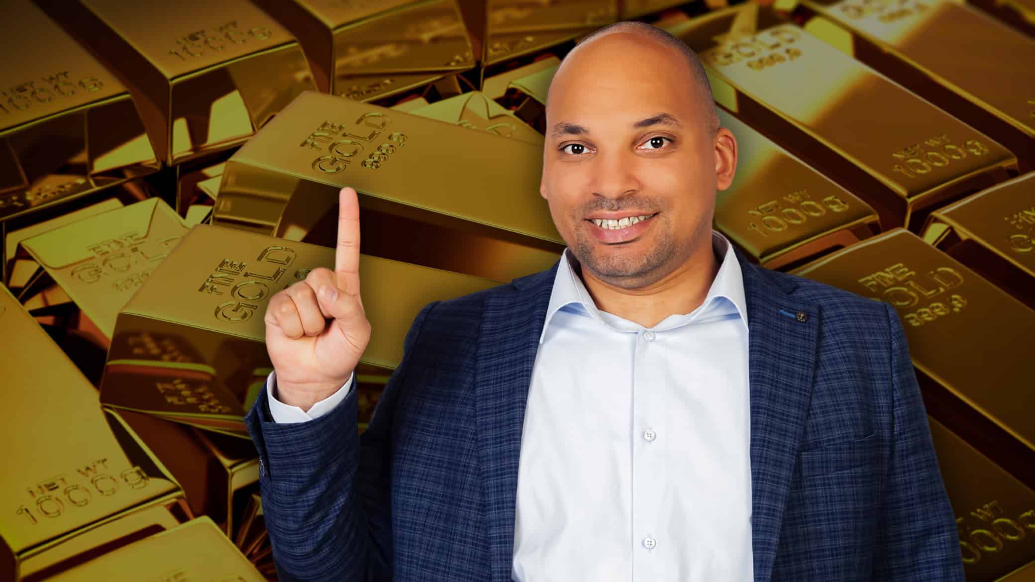A smiling bald man in a blue plaid suit jacket and light blue collared shirt points his right index finger upwards. He is positioned in the foreground against a blurred background of numerous stacked, shiny gold bars.