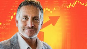 A middle-aged man with grey hair and a beard smiles confidently, set against a background of vibrant red, orange, and yellow. The background includes translucent yellow and white stock market candlestick charts, various numerical figures representing financial data, and a large orange arrow pointing diagonally upwards, symbolizing growth.
