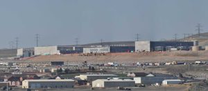 A wide, elevated view of a large industrial construction site on a clear day. In the middle ground, several large, gray, modern concrete buildings, typical of data centers, are under construction, with visible scaffolding and heavy machinery like bulldozers and excavators. Numerous electrical transmission towers with multiple lines line the barren hills in the background, while in the foreground, smaller residential or auxiliary buildings and roads are visible.