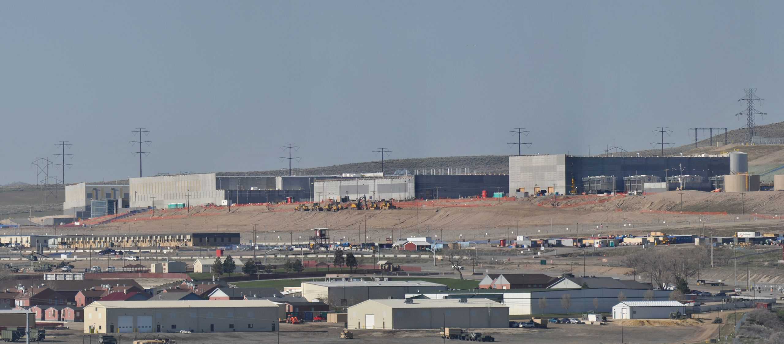 A wide, elevated view of a large industrial construction site on a clear day. In the middle ground, several large, gray, modern concrete buildings, typical of data centers, are under construction, with visible scaffolding and heavy machinery like bulldozers and excavators. Numerous electrical transmission towers with multiple lines line the barren hills in the background, while in the foreground, smaller residential or auxiliary buildings and roads are visible.