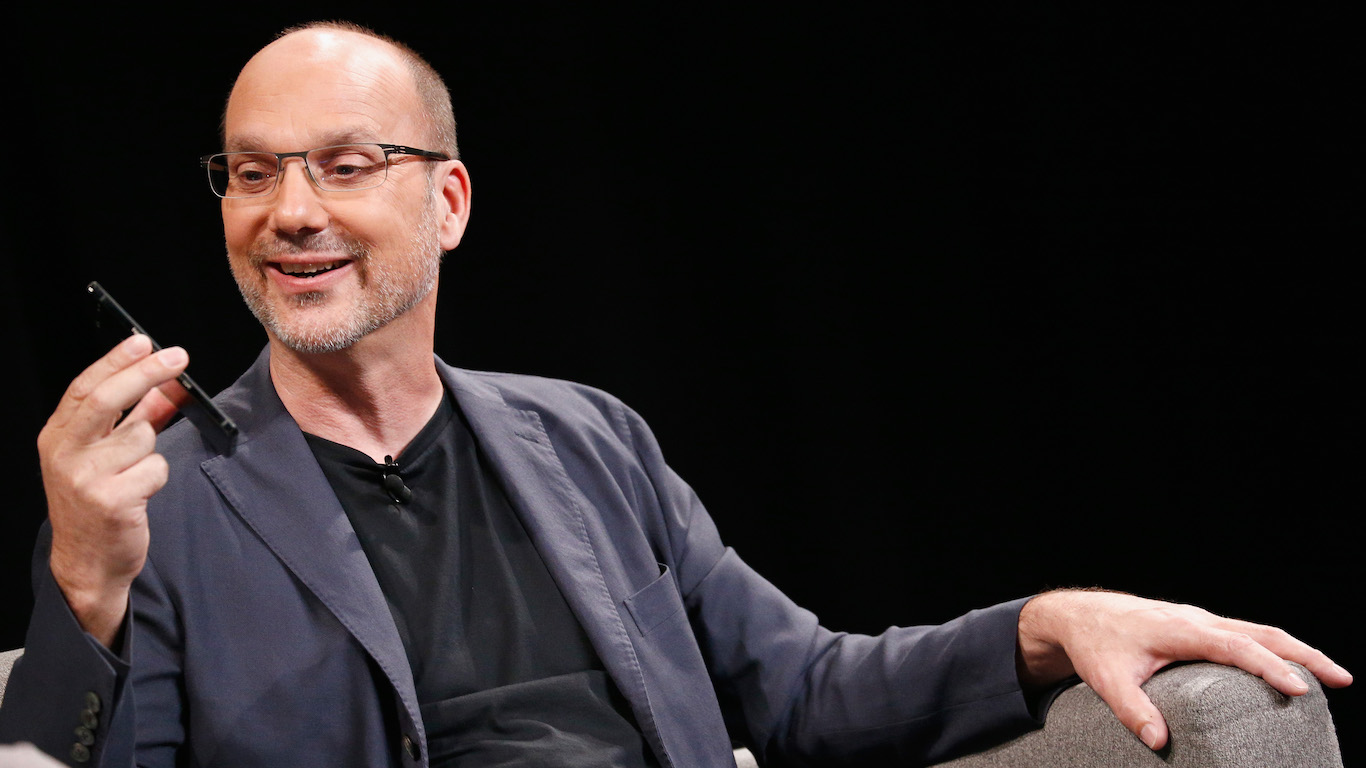 A man with short, balding hair and glasses, wearing a dark blue blazer and black t-shirt, smiles while holding a black smartphone in his left hand. He is looking down at the phone. His right arm rests on a grey upholstered chair. The background is a solid dark black.