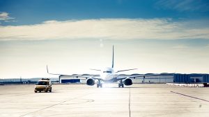 A wide shot of a white and blue commercial passenger jet, likely a Boeing 737, centered on a light gray concrete airport tarmac. The plane faces the viewer, with bright light or lens flare emanating from its nose and landing gear. A small yellow airport service vehicle is parked to the left. In the background, dark blue airport hangars are visible under a bright blue sky with scattered white clouds. The tarmac shows faint tire marks and linear markings.