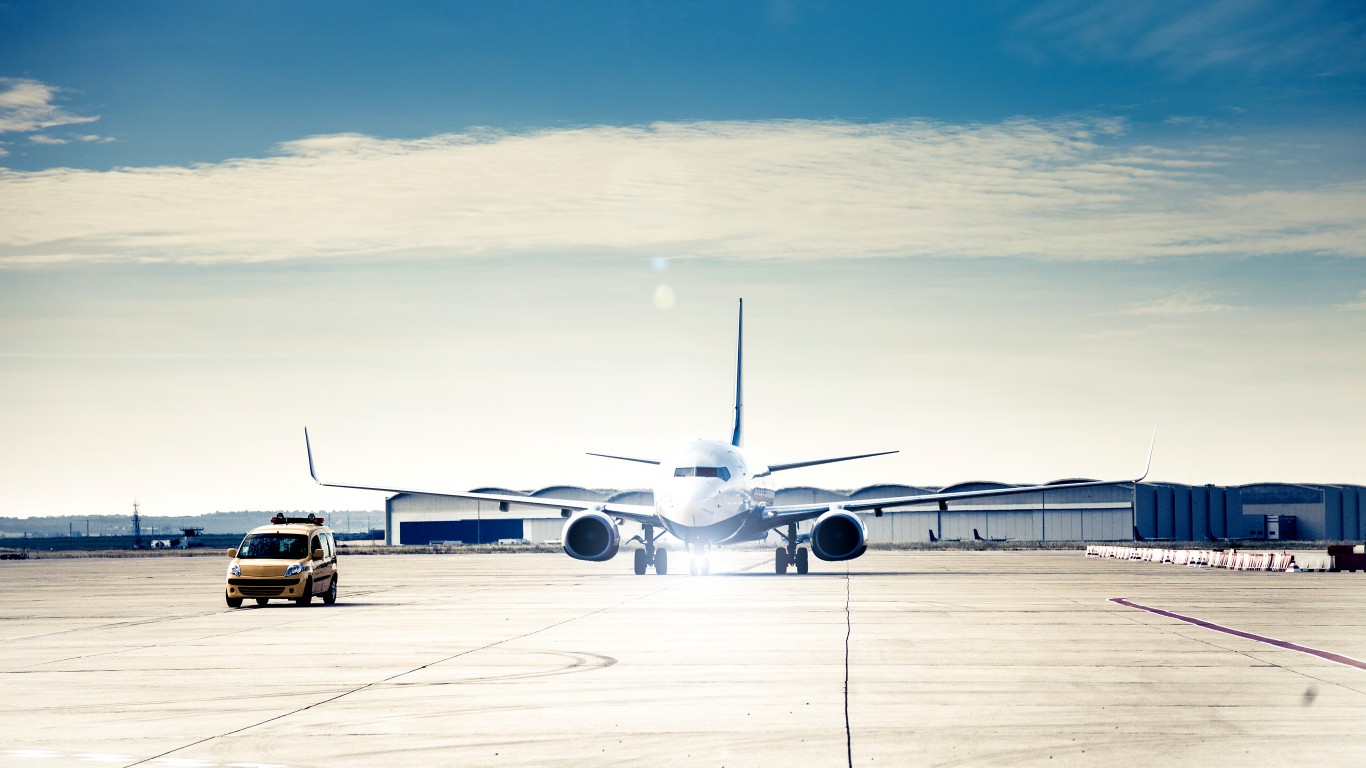 A wide shot of a white and blue commercial passenger jet, likely a Boeing 737, centered on a light gray concrete airport tarmac. The plane faces the viewer, with bright light or lens flare emanating from its nose and landing gear. A small yellow airport service vehicle is parked to the left. In the background, dark blue airport hangars are visible under a bright blue sky with scattered white clouds. The tarmac shows faint tire marks and linear markings.