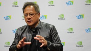 A man with short grey hair and glasses, wearing a black leather jacket over a black shirt, is shown from the chest up, gesturing with both hands while speaking. He has an earnest expression. The background is a white wall with repeating green and blue logos for NVIDIA and CES.