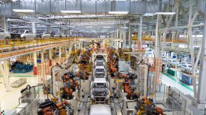 An overhead view inside a vast, modern car manufacturing factory. Silver car bodies move along assembly lines, surrounded by dozens of bright orange and yellow robotic arms performing welding and assembly tasks. Elevated conveyor belts with additional car chassis run in the background, beneath a high ceiling with extensive ductwork and lighting, conveying a sense of large-scale, automated industrial production.