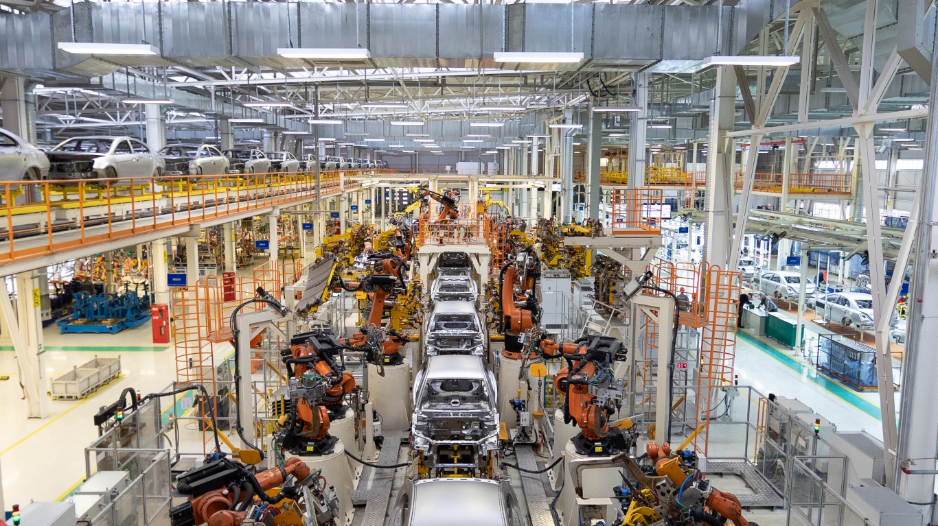 An overhead view inside a vast, modern car manufacturing factory. Silver car bodies move along assembly lines, surrounded by dozens of bright orange and yellow robotic arms performing welding and assembly tasks. Elevated conveyor belts with additional car chassis run in the background, beneath a high ceiling with extensive ductwork and lighting, conveying a sense of large-scale, automated industrial production.