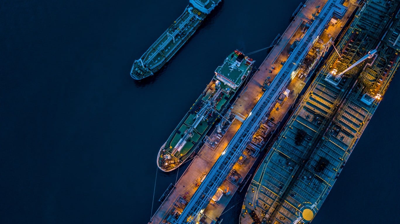 An aerial, high-angle view of multiple large oil tankers docked at an industrial port at night. The dark blue water surrounds the grey and teal ships, which are brightly illuminated by warm yellow lights from the pier and on their decks. Intricate piping structures are visible along the dock, connecting to the tankers, indicating active transfer operations. One tanker is further out in the dark water, while two are moored alongside the pier. The overall scene is dark with strong contrasts from the artificial lighting.