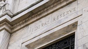 A close-up, low-angle view of the white marble exterior of the Federal Reserve building. The words 'FEDERAL RESERVE' are carved in capital letters into the stone facade, which exhibits subtle veins and a weathered appearance, with architectural cornices and molding details visible above the inscription.