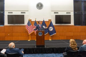 A man in a dark suit and red tie stands at a wooden podium, gesturing with open hands while speaking. Behind him on a stage are two American flags and two dark blue Virginia state flags. Above them, a large circular seal of Virginia is mounted on a white wall. Two large dark screens are also visible on the wall. In the foreground, the backs of several audience members are seen, including a man with gray hair wearing glasses and a woman with long blonde hair, attentively watching the speaker.