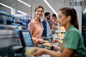 A smiling woman with long brown hair, wearing a peach-colored blouse, stands at a supermarket checkout counter. She holds a payment card as a cashier, whose face is out of focus, processes groceries including a loaf of bread and a packaged item. In the background, a man holding a child can be seen shopping in the bright, well-stocked aisles of the supermarket.
