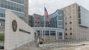 A wide-angle photograph of a large, modern United States Courthouse building. The structure features sections of light-colored stone, a prominent curved glass facade with multiple windows, and other parts with horizontal window accents. In the foreground, an American flag waves atop a tall flagpole. A stone wall on the left bears the words 'UNITED STATES COURTHOUSE' and displays the Great Seal of the United States.
