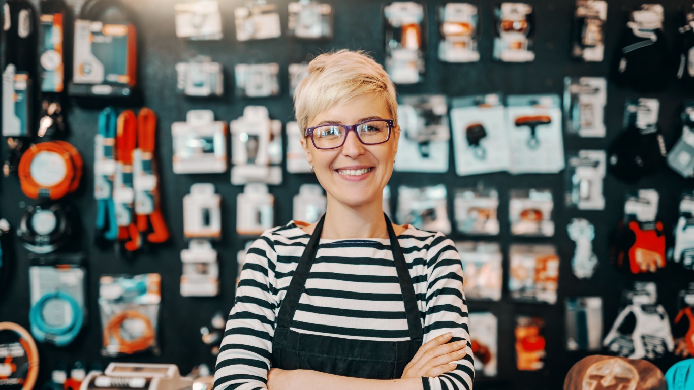 A smiling woman with short blonde hair and purple-framed glasses wears a black and white striped shirt and a black apron, with her arms crossed. She stands in a store with various packaged items, tools, and cables displayed on the dark wall behind her, which is softly blurred.