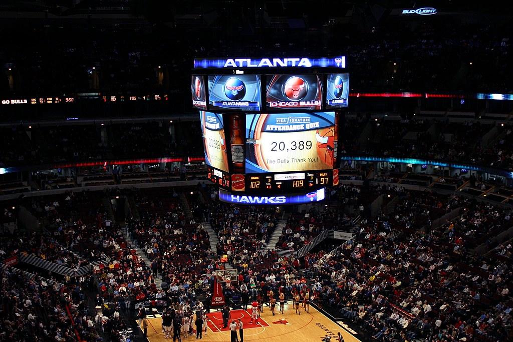 An aerial wide shot captures a dimly lit indoor basketball arena during an NBA game. A prominent, multi-sided video scoreboard hangs centrally, illuminated with blue and red graphics displaying scores for 'ATLANTA' and 'BULLS' (102 to 97), team logos for the Atlanta Hawks and Chicago Bulls, and an attendance count of 20,389. Below, the brightly lit wooden basketball court features players and officials, surrounded by a large crowd seated in the dark red and black tiered stands. The upper sections of the arena and a 'BUD LIGHT' sign are visible in the background.