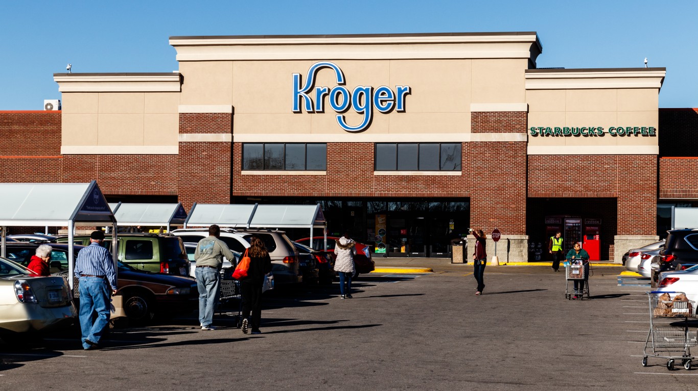 An outdoor shot of a Kroger supermarket building, featuring a prominent blue 'Kroger' logo on a beige facade above red brickwork. A 'Starbucks Coffee' sign is visible to the right. The foreground shows a busy parking lot with several cars, multiple shopping carts, and a number of people walking, some pushing carts, under a clear blue sky.