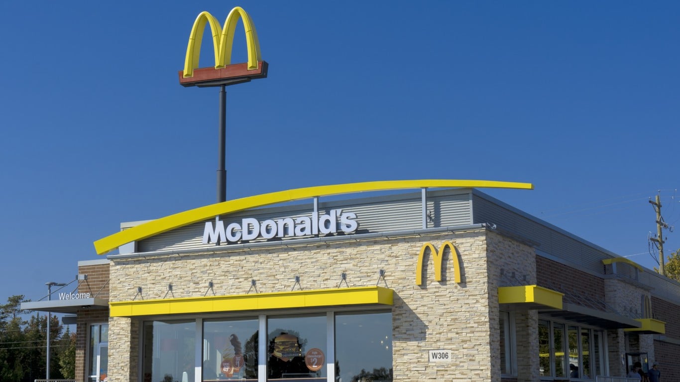 A clear blue sky above a modern McDonald's restaurant with a light-colored stone facade and large windows. A tall sign with the prominent golden arches 'M' logo stands on the left. The 'McDonald's' name is visible in white letters above the main entrance, and another golden 'M' logo is on the right side of the building. A 'Welcome' sign is visible near the entrance on the left, and a small sign with 'W306' is below a window on the right.