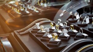 A close-up of a brightly lit jewelry display case filled with numerous sparkling luxury items. In the foreground, an assortment of diamond and gold rings are presented on individual black velvet stands. Behind them, several diamond necklaces and earrings are showcased on black busts and stands. The jewelry pieces are predominantly silver-toned and gold-toned, heavily encrusted with white gemstones that sparkle under the light. The background is softly blurred with warm, glowing reflections.