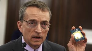 A man with short gray hair and glasses, wearing a dark suit and purple collared shirt, speaks into a microphone. He holds up a dark blue computer chip with a large, reflective golden-yellow square at its center in his left hand. The background is a blurred warm brown.