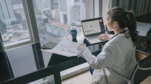 A person in a white lab coat with dark hair tied in a ponytail sits at a black desk, viewed from behind and to the side. They are looking at a silver laptop displaying colorful graphs and charts, while their left hand rests on an open white binder. A blue mug is on the desk. The desk is positioned in front of a large window offering a blurred view of a city skyline with tall buildings under a bright sky.