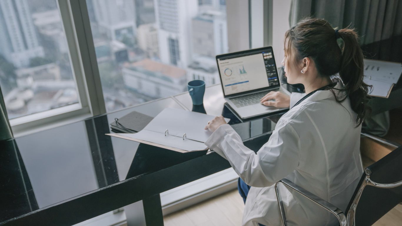 A person in a white lab coat with dark hair tied in a ponytail sits at a black desk, viewed from behind and to the side. They are looking at a silver laptop displaying colorful graphs and charts, while their left hand rests on an open white binder. A blue mug is on the desk. The desk is positioned in front of a large window offering a blurred view of a city skyline with tall buildings under a bright sky.