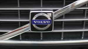 A close-up of a silver Volvo emblem with "VOLVO" in white on a dark blue rectangular background, encased in a larger silver circle with an arrow pointing right and up. The emblem is centered on a dark grey car grille with horizontal and vertical bars.