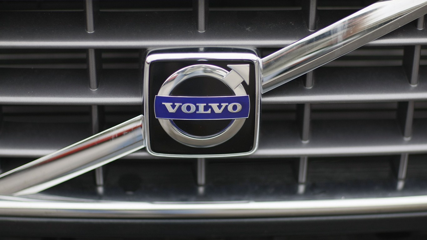 A close-up of a silver Volvo emblem with "VOLVO" in white on a dark blue rectangular background, encased in a larger silver circle with an arrow pointing right and up. The emblem is centered on a dark grey car grille with horizontal and vertical bars.