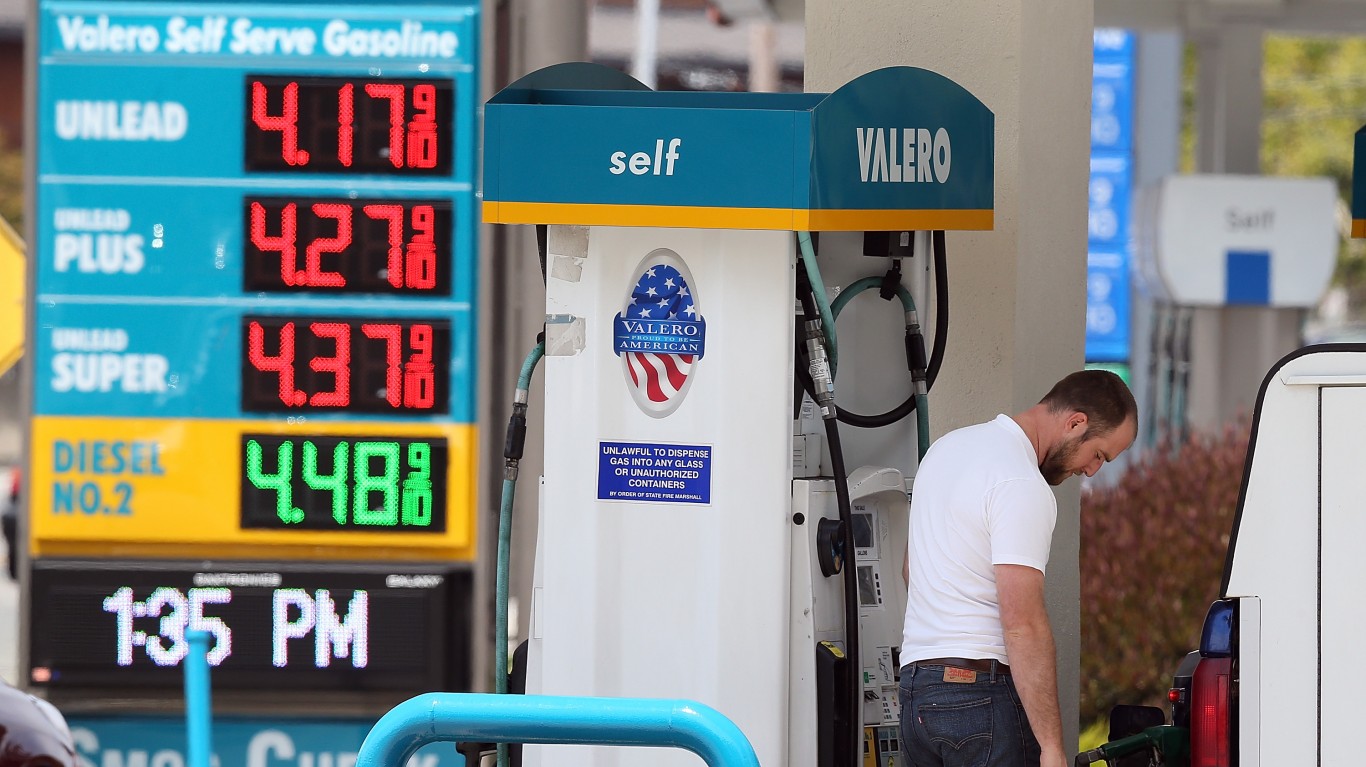 A man with a beard and a white t-shirt fills his car at a Valero self-serve gas station. To the left, a large blue and yellow digital sign displays gas prices: Unleaded $4.17 9/10, Unleaded Plus $4.27 9/10, Unleaded Super $4.37 9/10, and Diesel No. 2 $4.48 9/10, all in red and green numbers. The sign also shows '1:35 PM'. The gas pump itself is white with a 'self' sign, a Valero logo, and an American flag sticker.