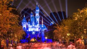 A long exposure photograph of Disneyland's Sleeping Beauty Castle at night, glowing in vibrant blue and white lights. Beams of blue and purple light fan out from behind the castle into the dark sky. A circular '60' emblem is prominently displayed on the castle's facade. In the foreground, a blurred crowd of people and trees with warm orange and yellow foliage create a dynamic and festive scene.