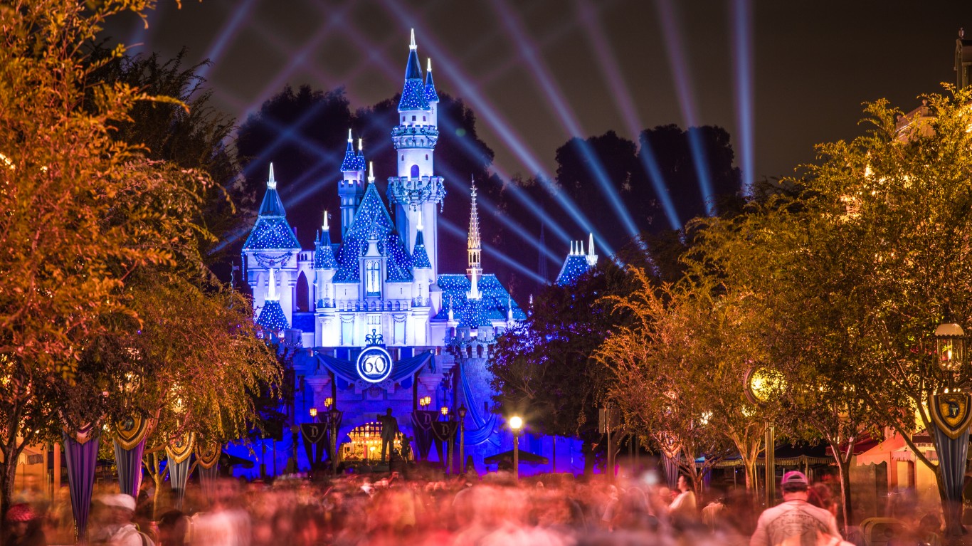 A long exposure photograph of Disneyland's Sleeping Beauty Castle at night, glowing in vibrant blue and white lights. Beams of blue and purple light fan out from behind the castle into the dark sky. A circular '60' emblem is prominently displayed on the castle's facade. In the foreground, a blurred crowd of people and trees with warm orange and yellow foliage create a dynamic and festive scene.
