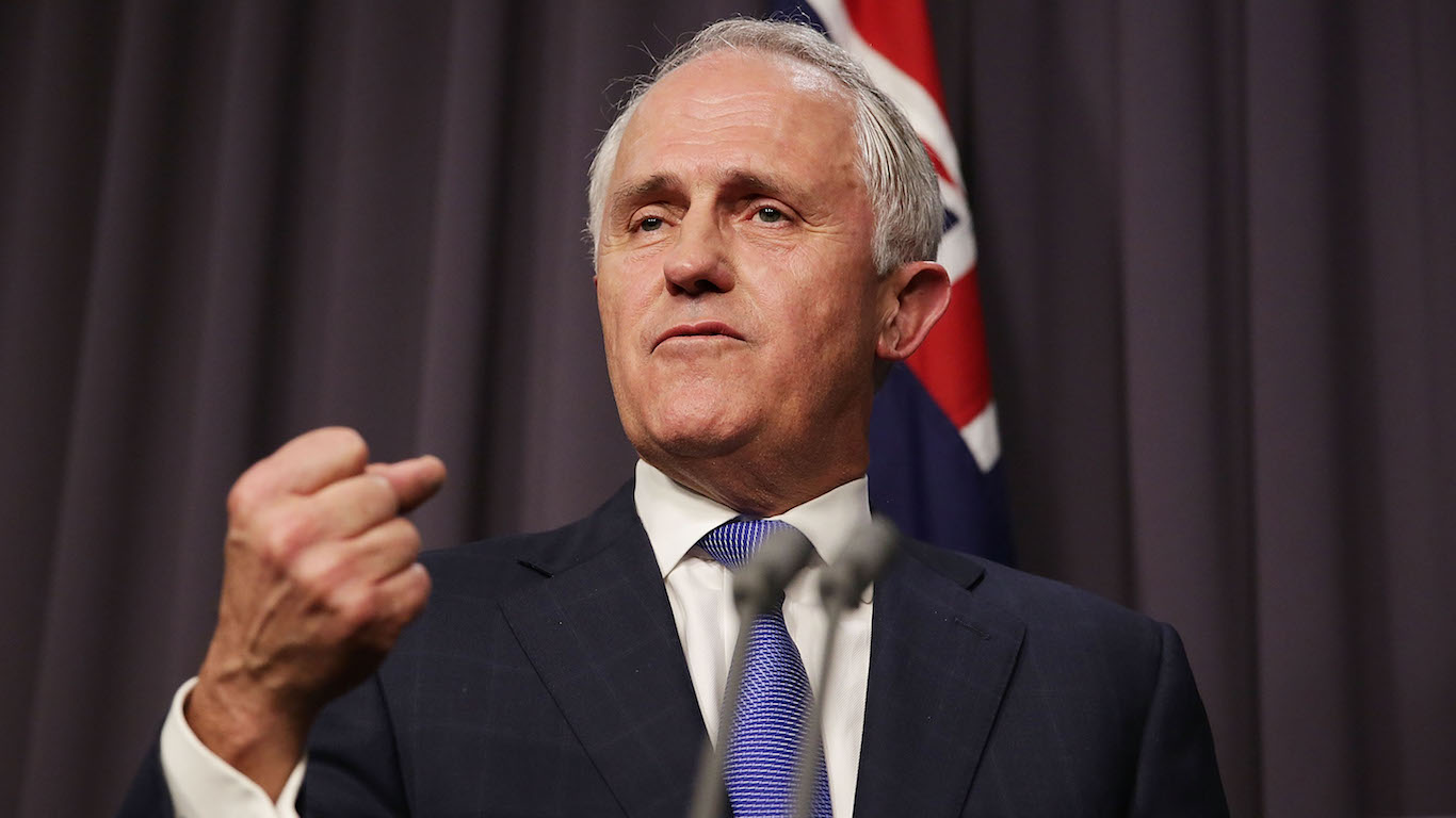 A man with gray hair, wearing a dark blue suit and a blue patterned tie, speaks assertively, with his right fist clenched and raised slightly. He stands in front of a dark curtain backdrop, with a corner of the Australian flag visible on his right side.