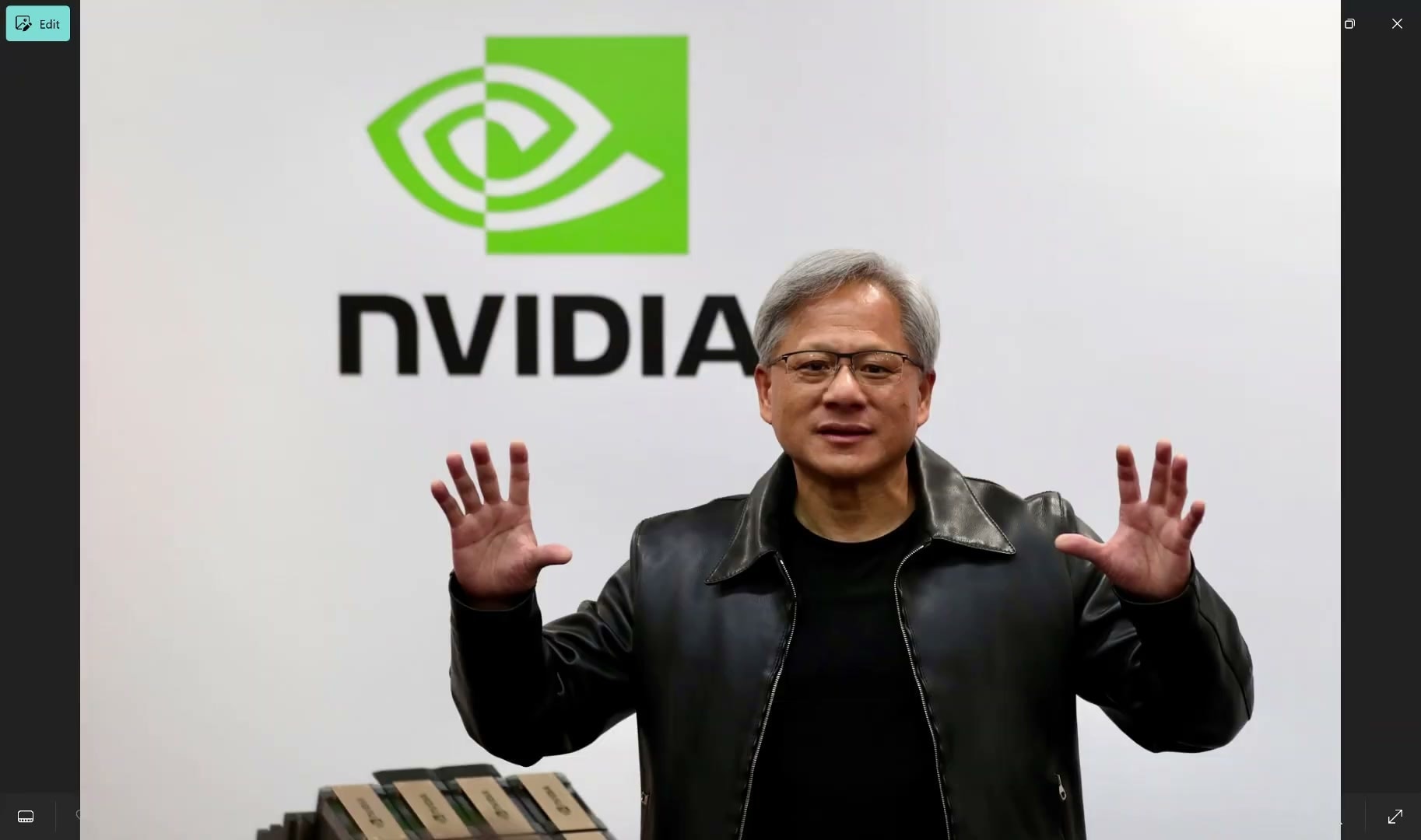 Jensen Huang stands in front of a white NVIDIA logo backdrop, wearing a black leather jacket, with both hands raised and open while speaking.