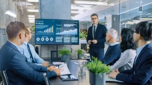 A male presenter in glasses and a dark sweater stands beside a large digital screen displaying financial analysis charts and graphs with a prominent "Analysis" title and a "+89%" indicator. He is addressing a group of five business professionals seated around a dark conference table in a modern office. The attendees are dressed in business attire and appear focused on the presentation. Potted green plants are visible on the table and around the room, which features glass partitions and modern lighting.
