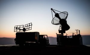 A stark silhouette of two large communication or radar antenna systems and a truck against a gradient sky. The sky transitions from light blue at the top to warm orange and pink hues at the horizon. The equipment on the left features a flat, grid-like antenna structure mounted on top of what appears to be a mobile command truck, while the right side shows a large, round parabolic dish antenna on a stationary base. The scene suggests a remote or strategic operational setting at dawn or dusk.