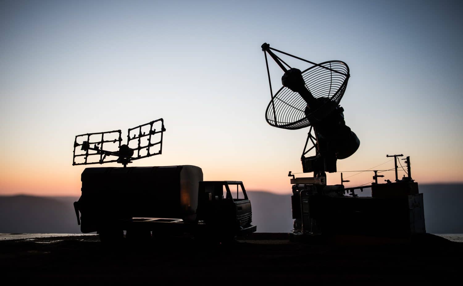 A stark silhouette of two large communication or radar antenna systems and a truck against a gradient sky. The sky transitions from light blue at the top to warm orange and pink hues at the horizon. The equipment on the left features a flat, grid-like antenna structure mounted on top of what appears to be a mobile command truck, while the right side shows a large, round parabolic dish antenna on a stationary base. The scene suggests a remote or strategic operational setting at dawn or dusk.