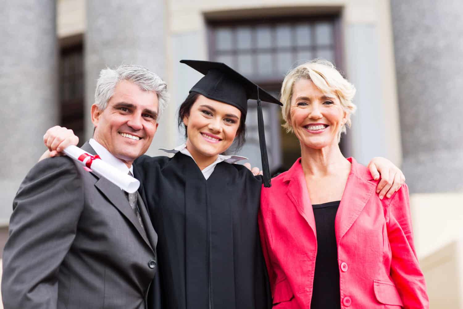 A young woman in a black graduation cap and gown stands between an older man in a dark suit and an older woman in a bright pink blazer. All three are smiling broadly at the viewer. The man on the left holds a white rolled-up diploma tied with a red ribbon. Their arms are around each other's shoulders, conveying a sense of pride and celebration.