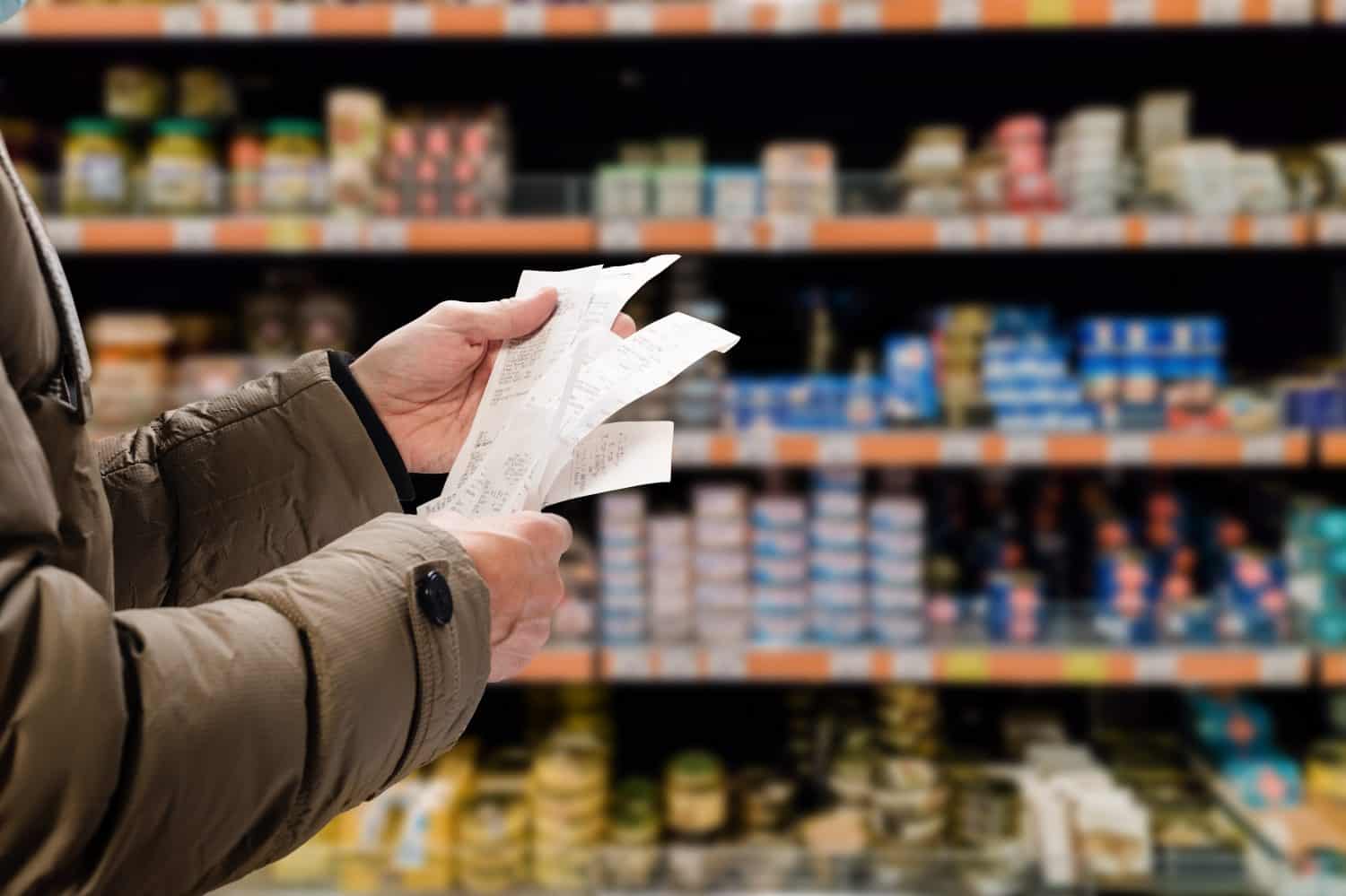 A close-up shot of a person's hands holding several crumpled white paper receipts. The person is wearing a brown winter jacket with a black cuff and button detail visible on the right sleeve. In the background, out-of-focus supermarket shelves are filled with various products, creating a blurred backdrop of colors and shapes.