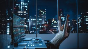 A man in a white shirt, leaning back in an office chair, raises both fists in a gesture of success. He is seated at a desk with multiple computer monitors displaying glowing financial charts and graphs, against a backdrop of a blurred, illuminated city skyline at night. The scene conveys a sense of achievement and late-night work in a modern office.