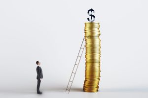 A businessman in a dark suit stands on the left, looking up at a very tall stack of gold coins on the right. A silver dollar sign sits on top of the coin stack. A wooden ladder leans against the stack of coins, reaching towards the top. The background is a clean, bright white.