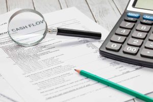 An overhead close-up view of financial documents on a light wooden desk. A magnifying glass rests over a section of text that prominently displays "CASH FLOW." A black calculator is positioned to the right of the document, and a green pencil lies across the lower part of the paper.