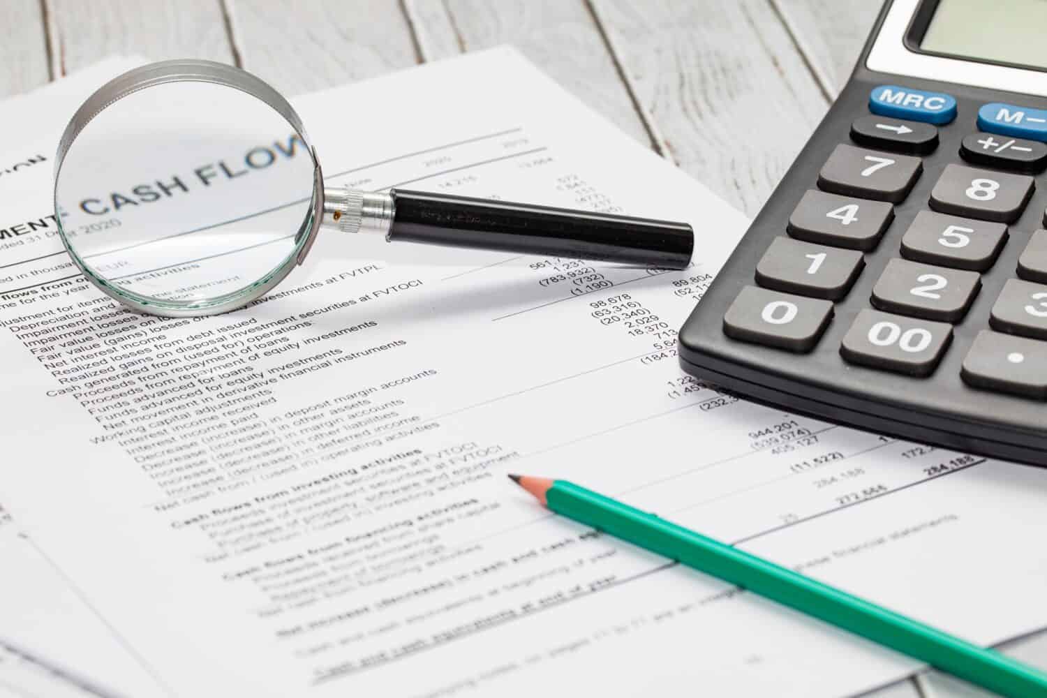 An overhead close-up view of financial documents on a light wooden desk. A magnifying glass rests over a section of text that prominently displays "CASH FLOW." A black calculator is positioned to the right of the document, and a green pencil lies across the lower part of the paper.