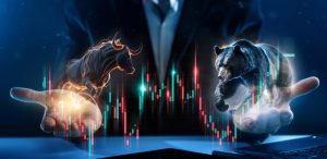 A stylized digital image shows two human hands emerging from a dark background, each holding a glowing animal. The left hand holds a powerful, golden-outlined bull, poised to charge upward. The right hand cradles a strong, blue-outlined bear, seemingly pushing downward. Between and around the hands are numerous vertical candlestick charts in vibrant red and teal, representing market data. A faint figure in a suit is visible in the background. The overall tone is dramatic and focused on financial market concepts.
