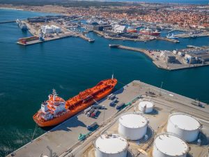 An aerial view captures a vibrant orange oil tanker docked at a busy commercial port. Next to the tanker, multiple white tanker trucks are lined up, along with several large, white cylindrical storage tanks on the pier. The expansive harbor includes various smaller vessels, cranes, and industrial structures, with a coastal town featuring red-roofed buildings sprawling into the distance under a bright sky.