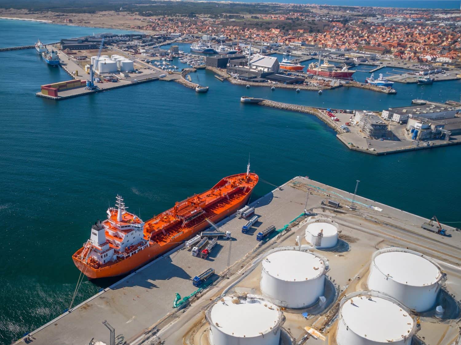 An aerial view captures a vibrant orange oil tanker docked at a busy commercial port. Next to the tanker, multiple white tanker trucks are lined up, along with several large, white cylindrical storage tanks on the pier. The expansive harbor includes various smaller vessels, cranes, and industrial structures, with a coastal town featuring red-roofed buildings sprawling into the distance under a bright sky.
